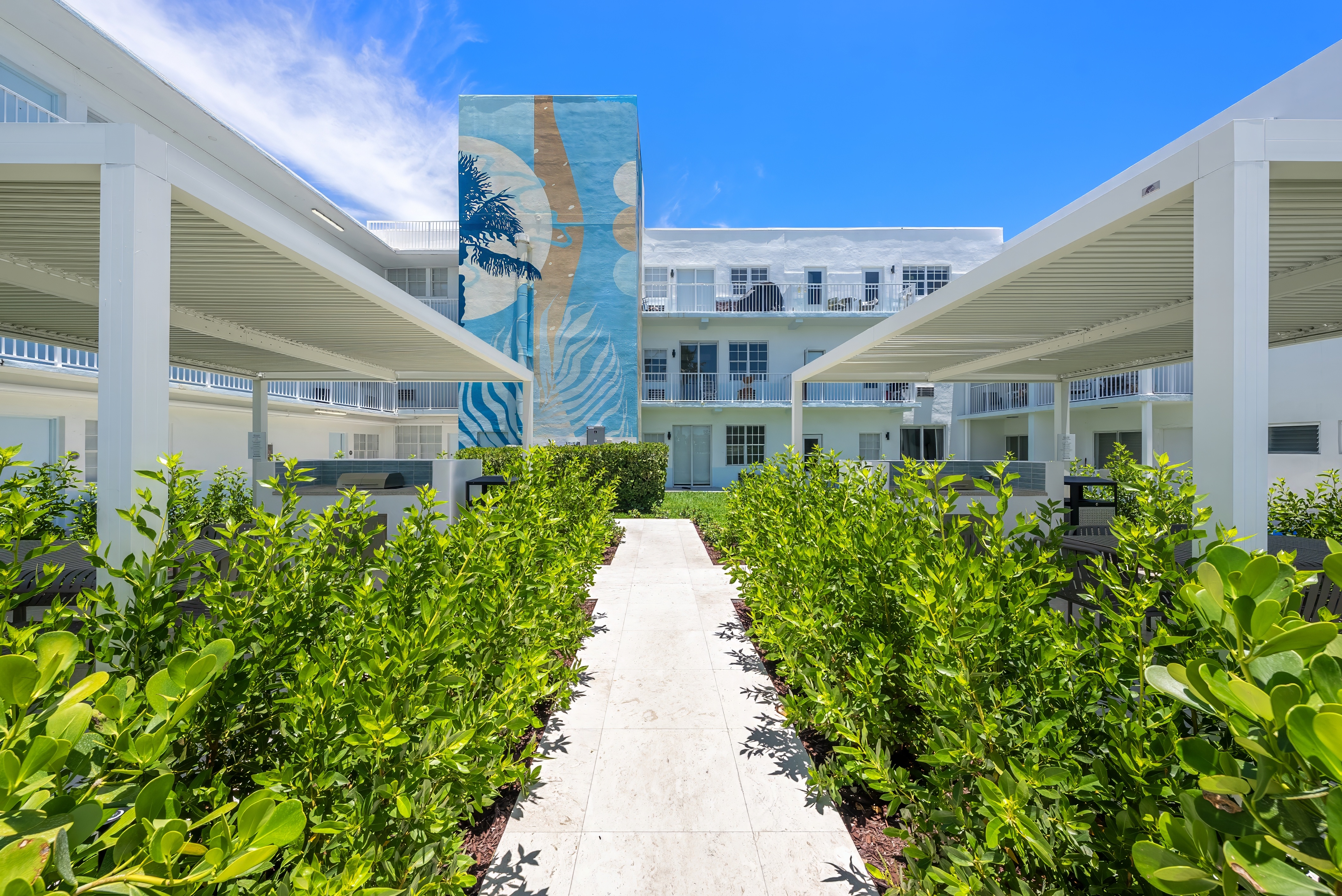 A long walkway with green plants on both sides leading to a building with a mural on it.